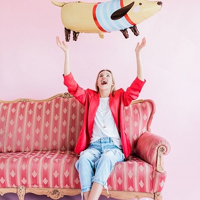 Mulher sentada num sofá vintage rosa a lançar balão em forma de cão castanho e azul.