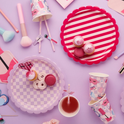 Pratos e copos de papel coloridos com cupcakes, macarons, pincéis de maquilhagem e bebida sobre fundo lilás