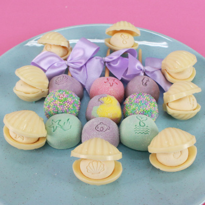 Doces coloridos em prato azul, conchas brancas e bolos redondos decorados.
