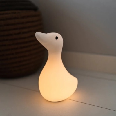 Abajur de mesa em forma de pato branco iluminado sobre chão branco junto a cesta rústica.