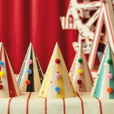 Chapéus de festa coloridos com pompons e estrelas numa mesa riscada com cortinas vermelhas ao fundo.