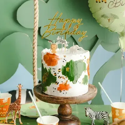 Bolo de aniversário com decoração de animais e topper Happy Birthday em suporte de madeira com balões e decoração temática de girafa.