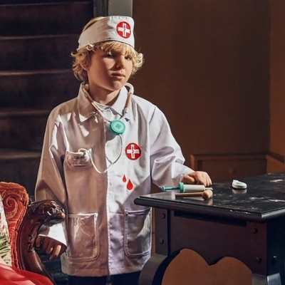 Criança vestida com jaleco branco de médico e gorro com cruz vermelha, em interior com mesa e brinquedos médicos.