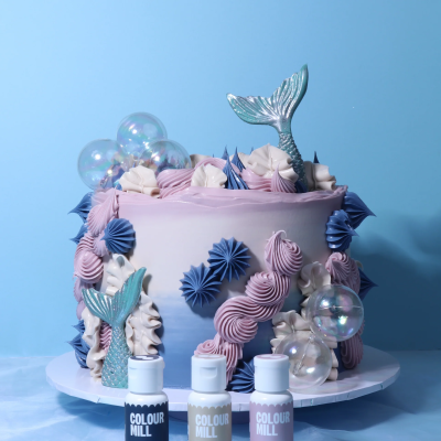 Bolo pastel decorado com cauda de sereia, ondas coloridas e bolhas, com frascos COLOUR MILL na base.