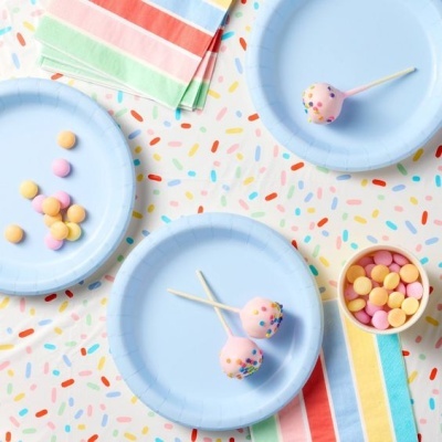 Pratos de papel azul com cake pops e confeitos coloridos sobre toalha e guardanapos coloridos