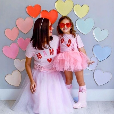 Mãe e filha com t-shirts cor de rosa e saias de tule, usando botas e óculos vermelhos, com fundo de corações coloridos.