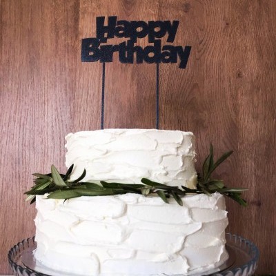 Bolo de aniversário de dois andares com creme branco, ramos verdes e topo de bolo 'Happy Birthday' em fundo de madeira