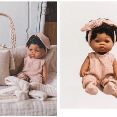 Boneca com macacão rosa e fita na cabeça com fundo branco e em cama bege