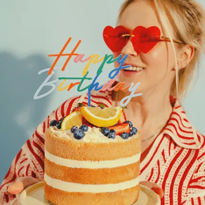 Bolo de aniversário com decoração de limão e mirtilos e topo colorido Happy Birthday, segurado por mulher com óculos de coração vermelhos