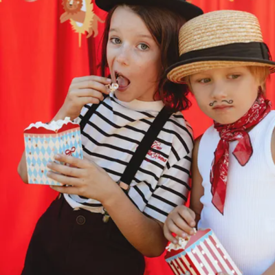 Duas crianças com roupas vintage segurando caixas de pipocas, com fundo vermelho e desenhos de animais