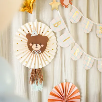 Decoração de festa infantil com painel de papel com desenho de urso, bandeirolas com letras BIRTH, estrelas douradas e decoração em leque