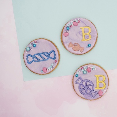 Três bolachas redondas decoradas com glacé lilás e desenhos coloridos em tecido