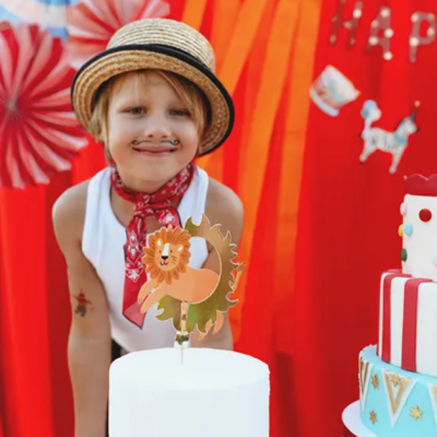 Criança com chapéu de palha e bigode desenhado atrás de bolo branco com decoração de leão