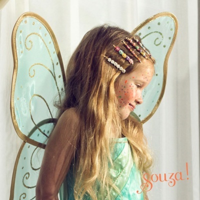 Criança com asas azuis e vestido verde, grampos de cabelo coloridos, fundo branco, texto 'souza!'