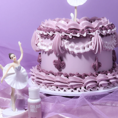 Bolo lilás decorado com glacê em ondas e laços, figura de bailarina e frasco de corante alimentar num fundo lilás com tecido.