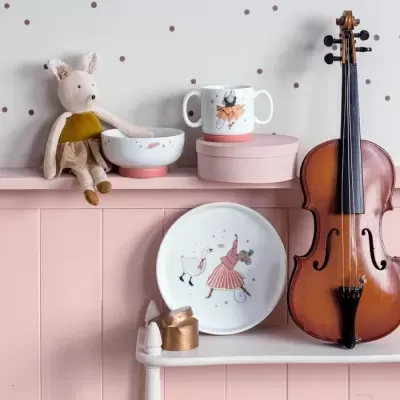 Conjunto de louça infantil com desenhos de animais, boneca de tecido, fita dourada e violino sobre móveis cor-de-rosa
