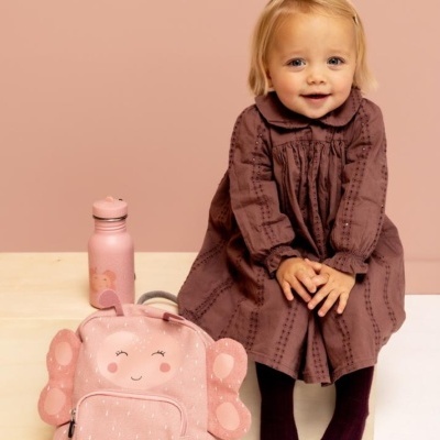 Criança com vestido castanho sentada ao lado de mochila rosa de asas de borboleta e garrafa térmica rosa
