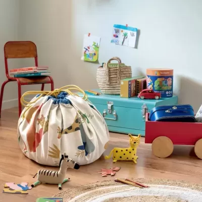 Saco redondo de tecido com desenhos de animais para arrumação de brinquedos infantis num quarto com brinquedos no chão e mala azul na carrinha vermelha.