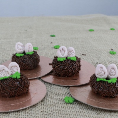 Quatro bombons com granulados de chocolate e ovos de açúcar decorativos em bases metálicas sobre tecido de juta