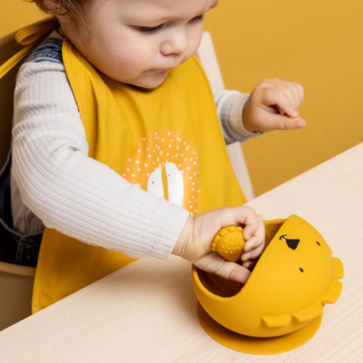 Copo de silicone amarelo em forma de leão com ventosa na base, usado por criança com babete amarelo sentado à mesa.