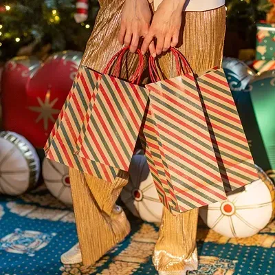 Sacos de papel riscado em verde, vermelho e bege segurados por pessoa com calças douradas, árvore de Natal ao fundo