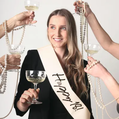 Mulher com faixa 'bride to be' e copos de champanhe rodeada por mãos com colares de pérolas