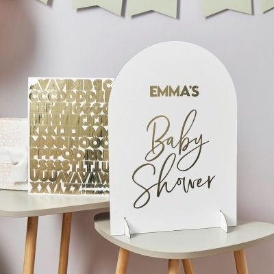 Placa decorativa branca com texto dourado EMMA'S Baby Shower em mesa