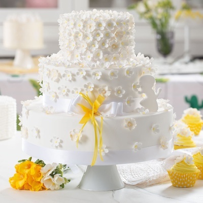 Bolo branco decorado com flores e laço amarelo num suporte branco