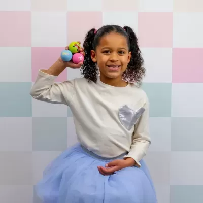 Menina com camisola creme, saia azul clara e bolas coloridas em fundo quadriculado pastel