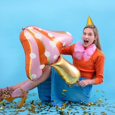 Mulher com balões metálicos rosa e dourado, vestida para festa, sentada em fundo azul claro com confetes