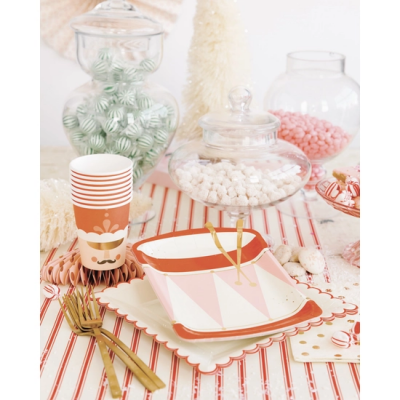 Mesa decorada com copos de papel, pratos coloridos, talheres dourados e frascos de vidro com doces.