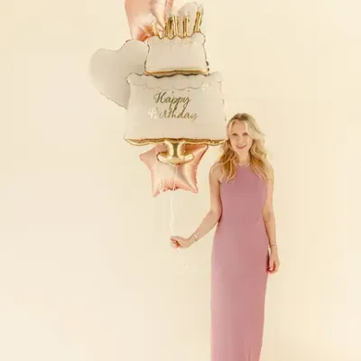 Mulher com vestido rosa segurando balão de aniversário em formato de bolo e estrelas