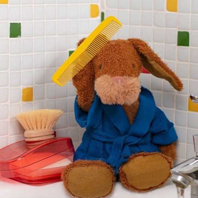 Urso de peluche com roupão azul numa casa de banho com escova e saboneteira