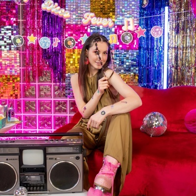 Mulher com roupa castanha, patins rosa, rádio grande prateado e fundo decorativo colorido com letreiro Disco Fever.