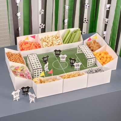 Tabuleiro de petiscos em forma de campo de futebol com vários snacks coloridos e figuras de jogadores