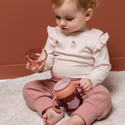 Bebé segurando copo infantil e recipiente cor de rosa sentado num tapete