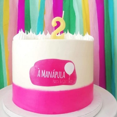 Bolo branco com base rosa, vela número 2 amarela e etiqueta rosa, com fundo colorido de fitas de papel