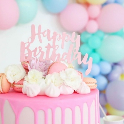 Bolo rosa decorado com macarons, flores e topper 'Happy Birthday' com balões coloridos ao fundo