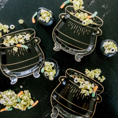 Pratos e copos de vidro preto decorados, com pipoca e gomas coloridas, em superfície preta.