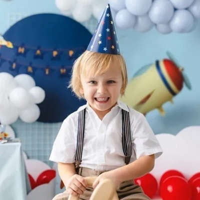 Criança loira com chapéu de festa azul e estrelas, segurando brinquedo de madeira, com balões coloridos ao fundo