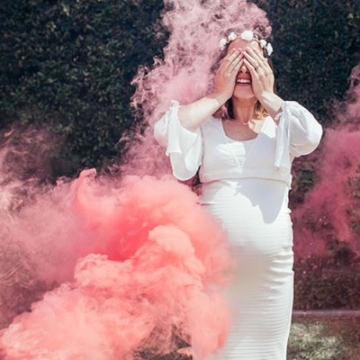 Pessoa com vestido branco e coroa de flores rodeada de fumo colorido ao ar livre