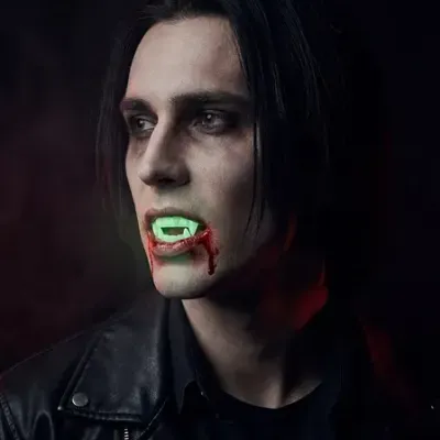 Homem com dentes de vampiro verdes e sangue artificial, vestindo casaco de couro preto em fundo escuro