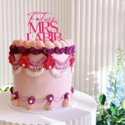 Bolo rosa decorado com creme roxo e rosa, texto Future MRS LABIR no topo, sobre prato branco junto a plantas