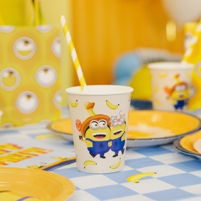Copo descartável branco com desenhos dos Minions e bananas com canudo amarelo e branco numa mesa com toalha azul e branca e pratos amarelos