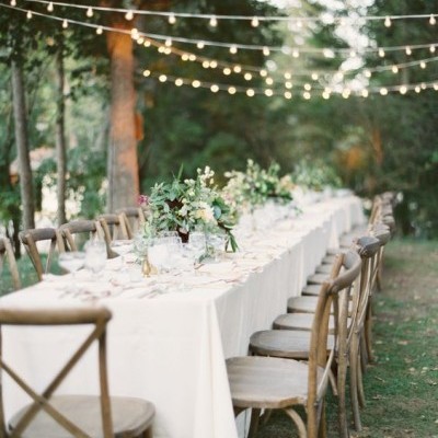 Mesa ao ar livre com toalha branca, cadeiras de madeira e decoração com flores e luzes suspensas