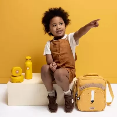 Criança com roupa castanha sentada ao lado de mochila e lanches amarelos em fundo amarelo.