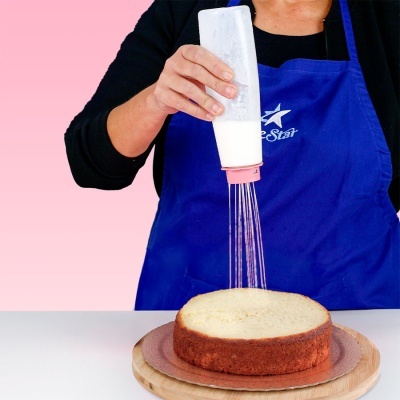 Pessoa a polvilhar açúcar em pó sobre um bolo redondo numa base de madeira.