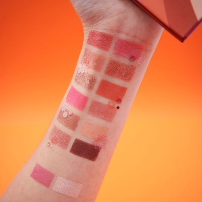 Amostras de maquilhagem em tons de rosa e castanho num braço com fundo laranja