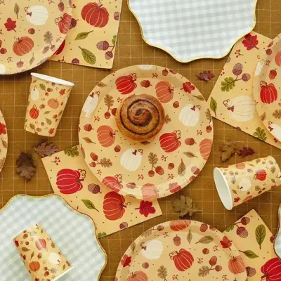 Artigos de festa descartáveis com padrão de abóboras e folhas outonais sobre mesa quadriculada castanha.