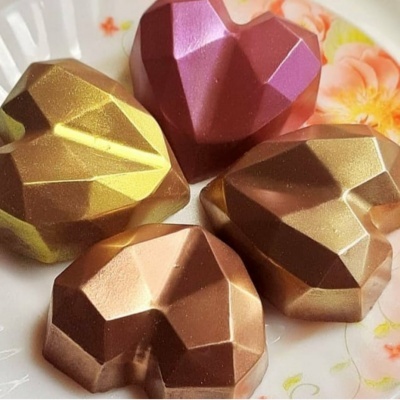 Quatro chocolates em forma de coração facetado em vários tons sobre uma chávena branca com flores.